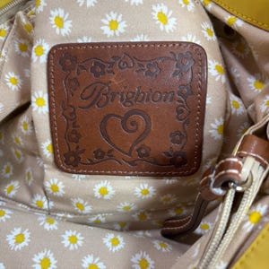 May include: Close-up of a yellow handbag interior. The interior lining features a daisy pattern on a beige background. A brown leather patch with the brand name "Brighton" is visible. The bag has a zipper and brown leather accents.