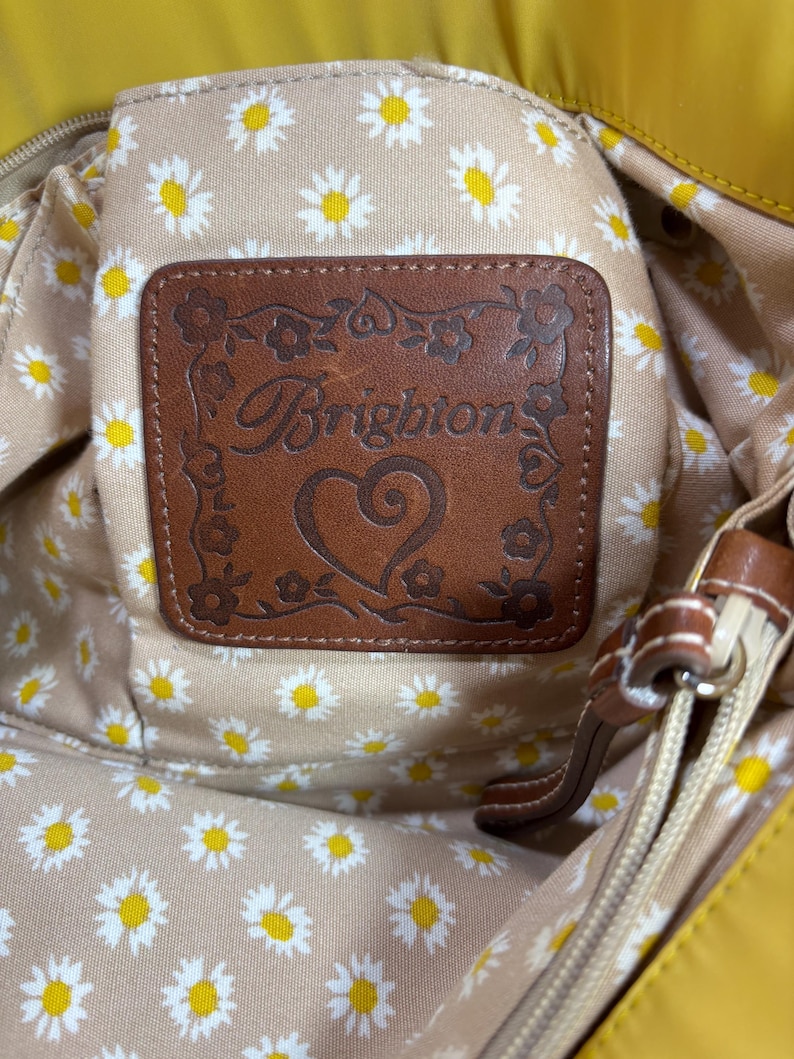 May include: Close-up of a yellow handbag interior. The interior lining features a daisy pattern on a beige background. A brown leather patch with the brand name "Brighton" is visible. The bag has a zipper and brown leather accents.