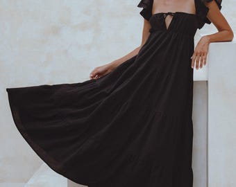 May include: A black maxi dress with a square neckline and ruffled cap sleeves. The dress features a tiered skirt and a keyhole detail at the bust. The model is wearing black sandals. The dress is made of a flowing fabric.