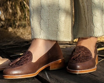 May include: Brown leather loafers with woven vamps and tassel details. The shoes have a low heel and are worn with cream-colored cable-knit pants. The shoes are on a wooden surface.