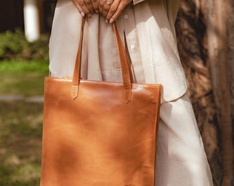 May include: A light brown leather tote bag with long handles. The bag is rectangular with a zipper closure and a small embossed logo on the lower front. The bag is held by a person wearing a beige outfit.