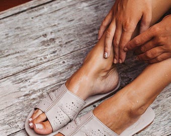 May include: A pair of light beige leather sandals with open toes and decorative cutouts. The sandals are resting on a weathered wooden surface. The image shows the sandals from a top-down perspective, highlighting the design and texture.