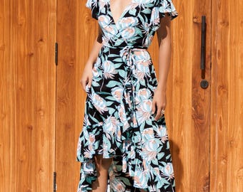 Puede incluir: Un vestido cruzado con escote en V y mangas cortas. El vestido presenta un fondo negro con un estampado floral en tonos azules, blancos y rosas. La falda tiene un dobladillo asimétrico. La modelo lleva sandalias negras.