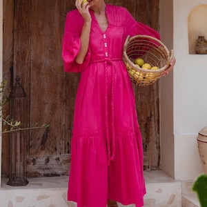 Pode incluir: Uma mulher em um vestido rosa vibrante segura uma cesta de limões. O vestido tem cintura marcada, mangas compridas e saia em camadas. Ela está em frente a uma porta de madeira.