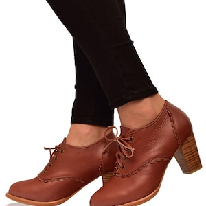 May include: Brown leather ankle boots with a scalloped edge design and a wooden block heel. The boots have a lace-up closure and are worn with black pants. The heels are approximately 3 inches high.