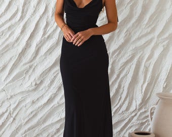 May include: A black maxi dress with a cowl neckline and spaghetti straps. The dress is floor-length and form-fitting, with a simple, elegant design. The model is wearing gold earrings and a bracelet. The background is a textured white wall.