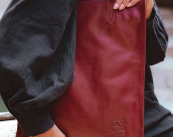 May include: A burgundy leather tote bag with a long, narrow design. The bag has a simple, elegant style with a single handle and a subtle embossed logo near the bottom. The bag is held by a person wearing a black long-sleeved shirt.