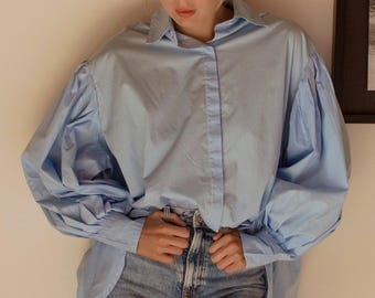 Könnte beinhalten: Ein hellblaues Button-Down-Hemd mit voluminösen Ärmeln und einem klassischen Kragen. Das Hemd wird mit hellblauen Jeans kombiniert. Der Gesamtstil ist lässig und entspannt, geeignet für den Alltag.