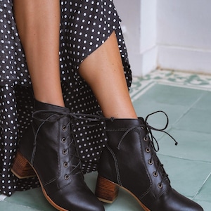 Könnte beinhalten: Schwarze Leder-Ankle Boots mit Schnürdesign und Holzblockabsätzen. Die Stiefel haben eine runde Spitze und eine hellbraune Sohle. Die Stiefel werden mit einem schwarz-weiß gepunkteten Kleid getragen.