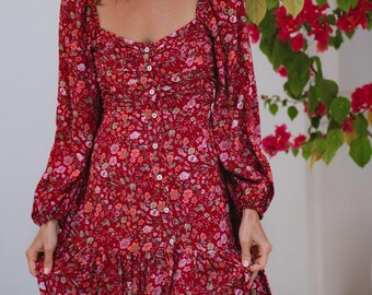 May include: A long-sleeved, floor-length dress in a deep red hue featuring an all-over floral pattern in shades of pink, orange, and white. The dress has a square neckline, a button-down front with small white buttons, and a gathered skirt.