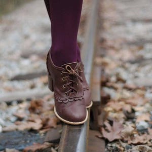 May include: A pair of brown leather shoes with scalloped edges and lace-up detailing. The shoes are worn with dark purple tights. The image is taken outdoors on a railroad track, with fallen leaves in the background.