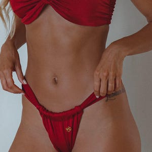 May include: A red bikini set. The top is a one-shoulder style with a knotted design. The bottom is a thong style with a small gold emblem. The fabric appears to be a smooth, solid color.