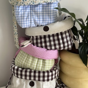 May include: A stack of fabric storage baskets featuring various patterns including blue and white gingham, brown and white gingham, light green gingham, and pink polka dots. Some baskets have floral linings and brown circular accents on the exterior.
