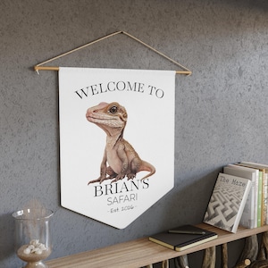 May include: A white banner with the text "WELCOME TO BRIAN'S SAFARI - Est 2026" and a detailed illustration of a lizard. The banner is suspended from a wooden rod with a string.