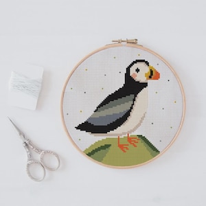 Könnte beinhalten: Kreuzstich-Stickerei eines Papageientauchers in einem Holzreifen. Der Vogel hat schwarze, weiße und graue Federn, einen orangefarbenen Schnabel und orangefarbene Füße. Der Hintergrund ist weiß mit kleinen grünen und gelben Punkten. Schere und Faden liegen neben dem Reifen.