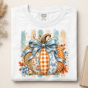 May include: A white t-shirt featuring a graphic of a pumpkin decorated with a large blue bow. The pumpkin segments display various patterns including leopard print, orange gingham, floral motifs, and polka dots, surrounded by autumn leaves.