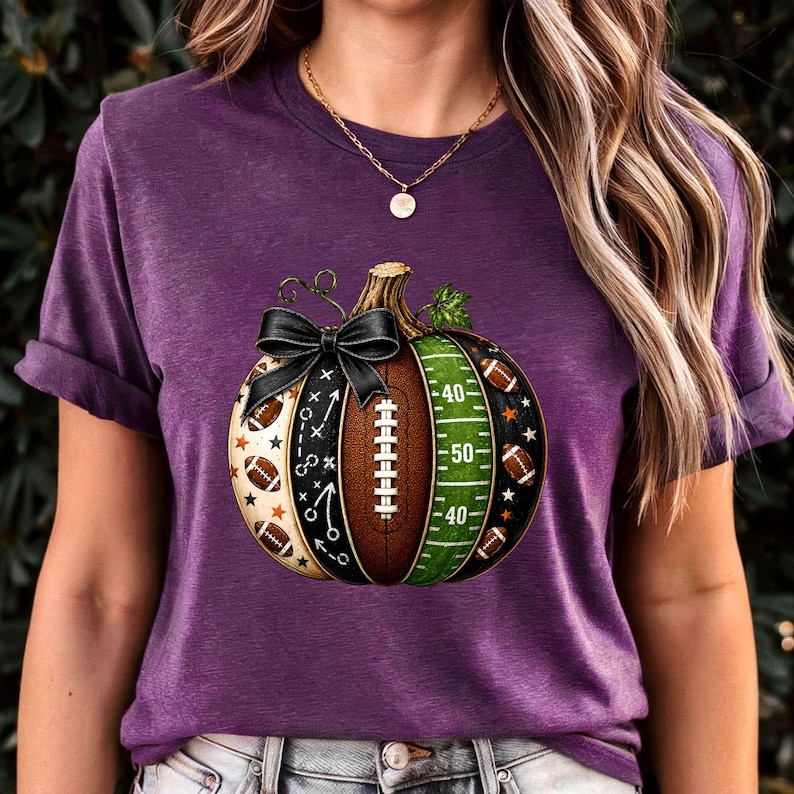 May include: A purple short-sleeve t-shirt featuring a graphic of a pumpkin designed with football-themed elements. The pumpkin includes a black bow, a brown football texture, a green field pattern with yard lines, and small football icons.