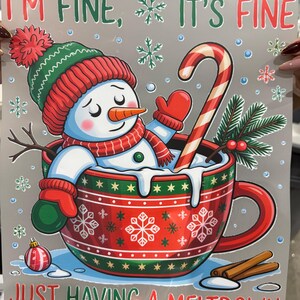 Może przedstawiać: Ilustracja przedstawia bałwana w czerwonym kubku z napisem "I'M FINE, IT'S FINE, JUST HAVING A MELTDOWN." Bałwanek ma na sobie zielono-czerwoną czapkę, szalik i rękawiczki. W kubku znajduje się r&oacute;wnież laska cukrowa i laski cynamonu.