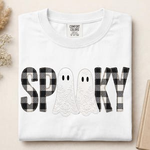 May include: A white crew neck t-shirt featuring the text SPOOKY printed across the chest. The letters S, P, K, and Y are filled with a black and white buffalo plaid pattern, while the two O's are replaced by white ghost shapes with floral lace details.