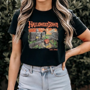 May include: Black t-shirt featuring a Halloween Town graphic. The design includes a pumpkin, buildings, and the text "HALLOWEENTOWN" in orange, with "EST. 1998" below. The shirt is a classic crew neck.