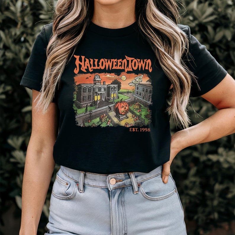 May include: Black t-shirt featuring a Halloween Town graphic. The design includes a pumpkin, buildings, and the text "HALLOWEENTOWN" in orange, with "EST. 1998" below. The shirt is a classic crew neck.