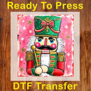 May include: A colorful DTF transfer featuring a whimsical nutcracker soldier. The design showcases a green hat with a red bow, a red and green uniform, and a gold scepter. The background is pink with white stars and the text "Ready To Press".
