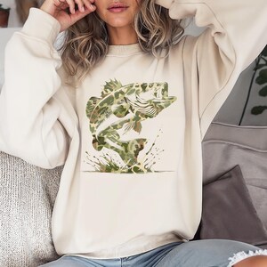 Puede incluir: Sudadera de color crema con un dise&ntilde;o de pez lubina saltando en patr&oacute;n de camuflaje. El pez se representa con la boca abierta y detalles de salpicaduras de agua. La sudadera es de estilo casual.