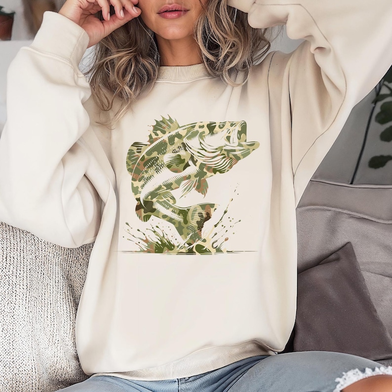 Puede incluir: Sudadera de color crema con un dise&ntilde;o de pez lubina saltando en patr&oacute;n de camuflaje. El pez se representa con la boca abierta y detalles de salpicaduras de agua. La sudadera es de estilo casual.