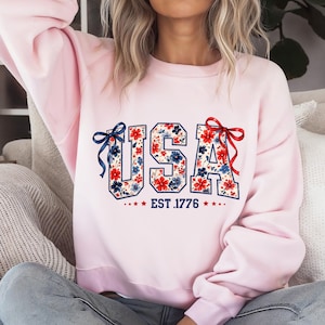 Może przedstawiać: Jasnor&oacute;żowa bluza z napisem "USA" w kwiatowy wz&oacute;r z czerwonych i niebieskich kwiat&oacute;w. Pod napisem "USA" znajduje się napis "EST. 1776". Bluza ma czerwoną kokardę po prawej stronie i niebieską kokardę po lewej stronie.