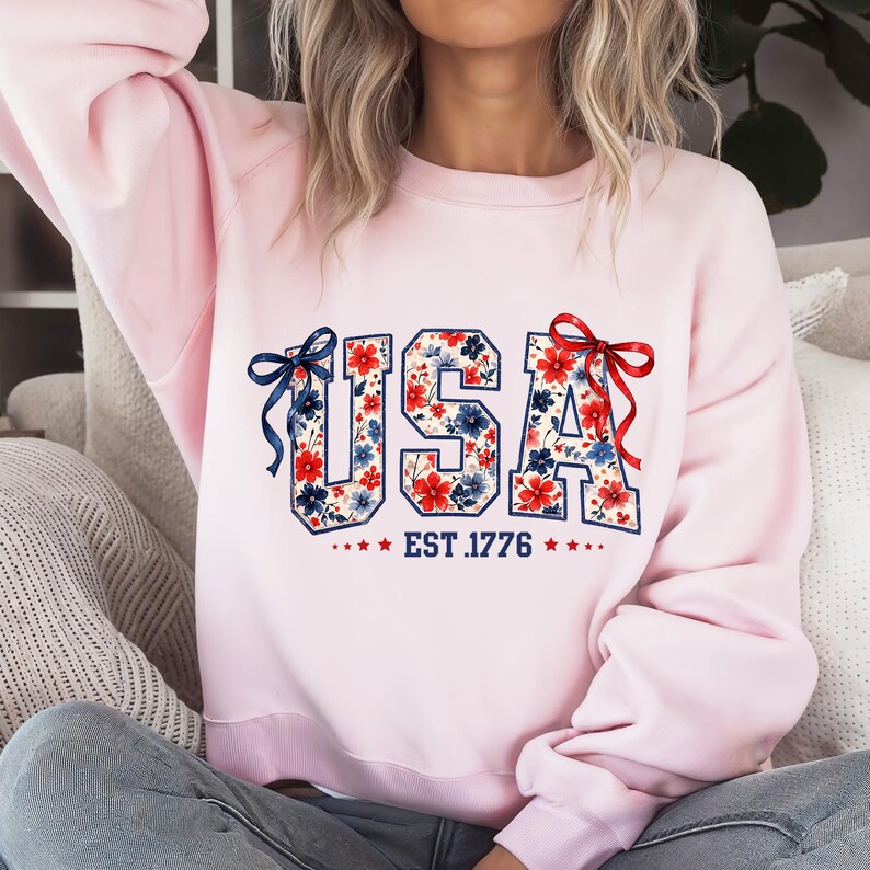 Może przedstawiać: Jasnor&oacute;żowa bluza z napisem "USA" w kwiatowy wz&oacute;r z czerwonych i niebieskich kwiat&oacute;w. Pod napisem "USA" znajduje się napis "EST. 1776". Bluza ma czerwoną kokardę po prawej stronie i niebieską kokardę po lewej stronie.