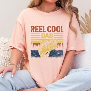 May include: A light peach-colored t-shirt with the text "REEL COOL DAD" in a distressed, vintage style. Below the text is a graphic of a fish jumping out of the water, with a retro sunset design. The shirt is a relaxed fit.