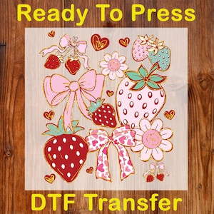 May include: A DTF transfer sheet featuring a strawberry theme. The design includes strawberries, bows, and flowers in pink, red, and gold. The text "Ready To Press" and "DTF Transfer" are in yellow.
