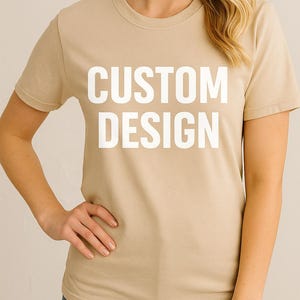 May include: A tan-colored t-shirt with the words "CUSTOM DESIGN" printed in large, white capital letters. The shirt has short sleeves and a crew neck. The model is wearing blue jeans.