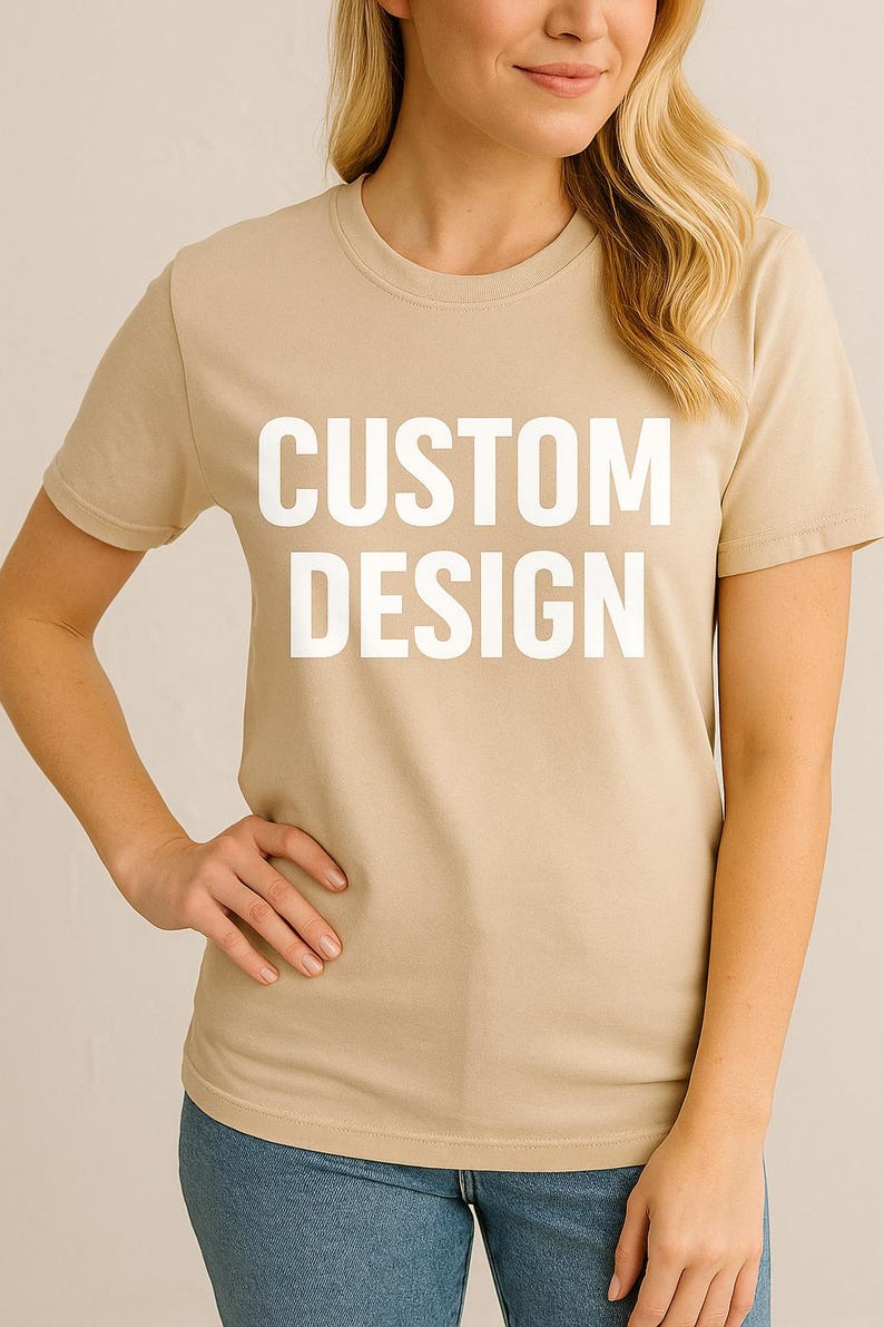 May include: A tan-colored t-shirt with the words "CUSTOM DESIGN" printed in large, white capital letters. The shirt has short sleeves and a crew neck. The model is wearing blue jeans.