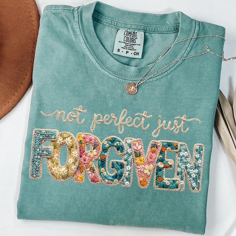 Op de afbeelding: Een turquoise Comfort Colors T-shirt met de geborduurde tekst "not perfect just FORGIVEN". Het woord "FORGIVEN" heeft een bloemenmotief. Een zilveren ketting met een kleine hanger ligt op het shirt.