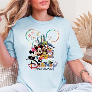 May include: Pale blue t-shirt with a vibrant Disney-themed graphic. The design showcases Mickey and Minnie Mouse, Donald Duck, and other characters, with the words "Disney Family Vacation" and the year "2026".