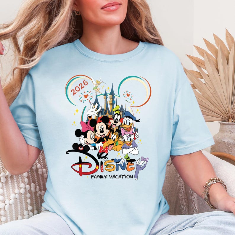 May include: Pale blue t-shirt with a vibrant Disney-themed graphic. The design showcases Mickey and Minnie Mouse, Donald Duck, and other characters, with the words "Disney Family Vacation" and the year "2026".