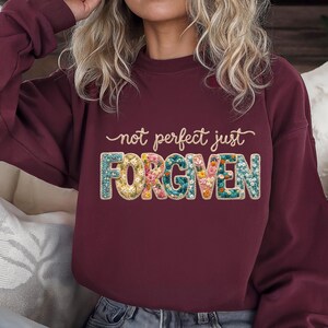Op de afbeelding: Bordeaux sweatshirt met de tekst "not perfect just FORGIVEN" in een bloemenontwerp. De sweatshirt heeft een effen bordeauxrode kleur. De tekst is in een scriptlettertype met het woord "FORGIVEN" in een bloemenontwerp.