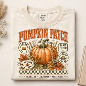 May include: A cream-colored t-shirt featuring a large orange pumpkin graphic with the text PUMPKIN PATCH. The design includes smaller circular labels reading FRESH FROM THE PATCH and LOCALLY GROWN, plus a tag labeled AUTUMN PICKED.