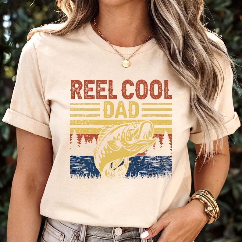 May include: Cream-colored t-shirt with the text "REEL COOL DAD" in a distressed, vintage style. The design includes a graphic of a leaping fish, set against a backdrop of horizontal stripes in yellow, red, and blue, evoking a sunset or sunrise.