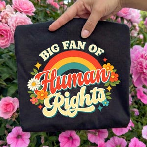 May include: Black t-shirt with the text "BIG FAN OF Human Rights" in a retro design. The design includes a rainbow, flowers, and stars. The shirt is held up by a hand in front of a background of pink flowers.