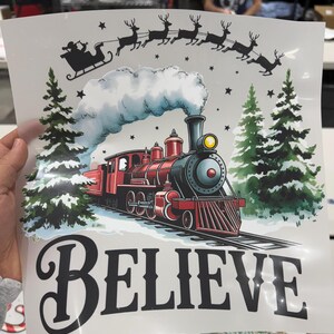 Pu&ograve; includere: Grafica a tema natalizio con un treno a vapore rosso sui binari, la slitta di Babbo Natale e le renne sopra. La parola &laquo;BELIEVE&raquo; &egrave; visualizzata in grassetto, in nero. Pini innevati e stelle completano il design festivo.