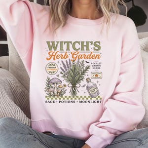 May include: A light pink crewneck sweatshirt featuring a graphic print with the text "WITCH'S Herb Garden" and "SAGE &bull; POTIONS &bull; MOONLIGHT". The design includes illustrations of a bundle of herbs, mushrooms, a potion bottle, and small stars.