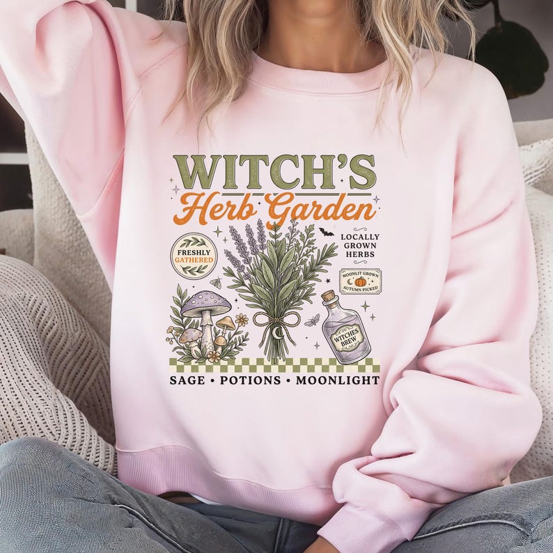 May include: A light pink crewneck sweatshirt featuring a graphic print with the text "WITCH'S Herb Garden" and "SAGE &bull; POTIONS &bull; MOONLIGHT". The design includes illustrations of a bundle of herbs, mushrooms, a potion bottle, and small stars.