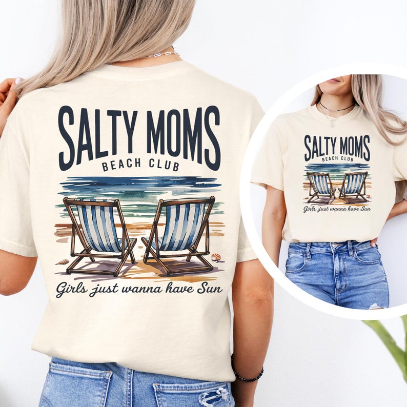 May include: Cream-coloured t-shirt with a beach-themed graphic. The design features the text "SALTY MOMS BEACH CLUB" above a watercolour illustration of a beach scene with two beach chairs. The phrase "Girls just wanna have Sun" is printed below.
