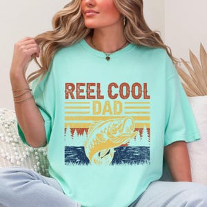May include: A light teal t-shirt with the text "REEL COOL DAD" in brown and yellow. Below the text is a graphic of a fish jumping out of the water, with a blue and yellow striped background. The shirt is a comfortable fit.