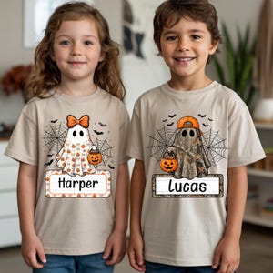 May include: Two beige t-shirts featuring Halloween graphics. The left shirt shows a ghost with an orange bow and pumpkin bucket above a name tag reading Harper. The right shirt shows a ghost in a camo print with an orange cap and pumpkin bucket above a name tag reading Lucas. Both designs include spiderwebs and bats.