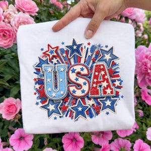 May include: White sweatshirt with a patriotic USA design. The design features the letters "USA" in red, white, and blue, surrounded by stars and bursts. The sweatshirt is held up by a hand, with a background of pink flowers.