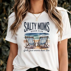 May include: White t-shirt featuring the text "SALTY MOMS BEACH CLUB" above a beach scene with two deckchairs. The phrase "Girls just wanna have Sun" is printed below the chairs.