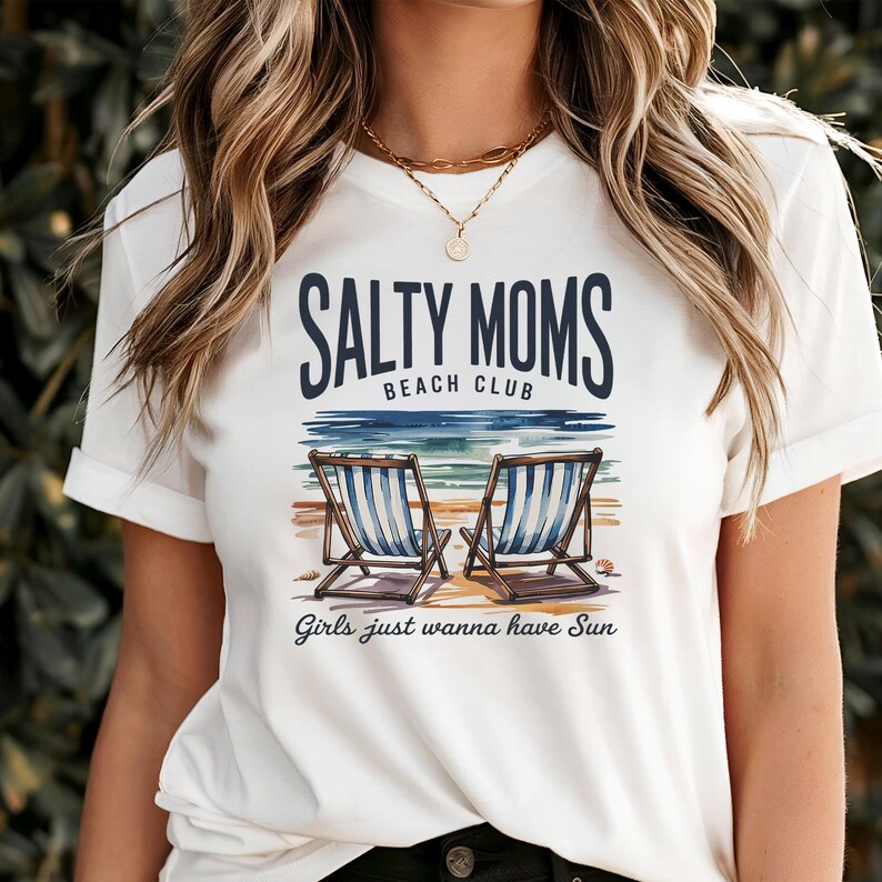 May include: White t-shirt featuring the text "SALTY MOMS BEACH CLUB" above a beach scene with two deckchairs. The phrase "Girls just wanna have Sun" is printed below the chairs.